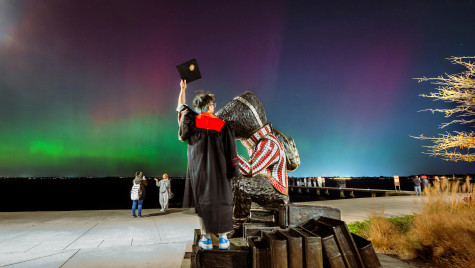 A student wearing a graduation gown stands next to a Bucky Badger statue while holding his cap to the sky. In front of him, Northern Lights dance across the sky.