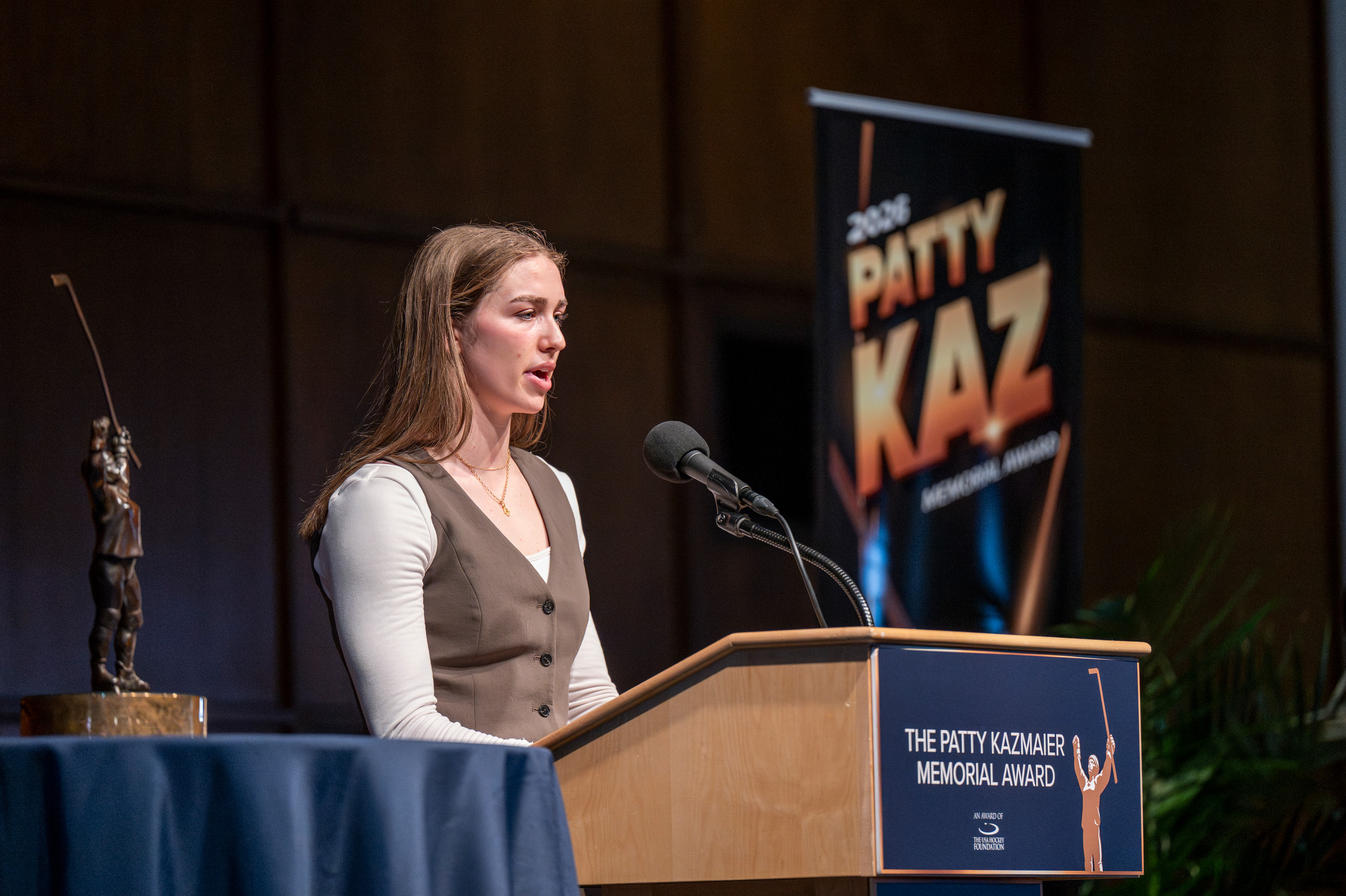 Harvey stands on stage behind a wooden podium and addresses a crowd. Next to her is a tablecloth-covered stand that holds a gold trophy.