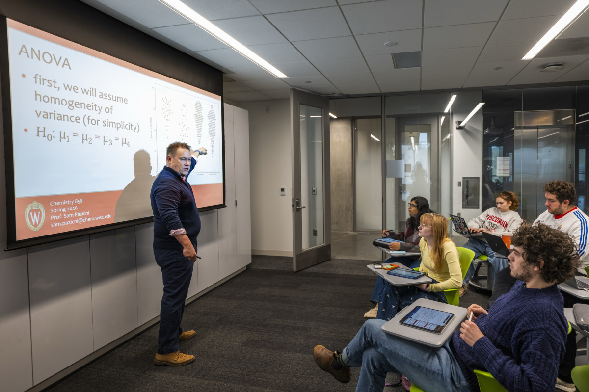Sam Pazicni stands at the head of a classroom and points to a data set on the projector screen. He is addressing a small group of students sitting in front of him.