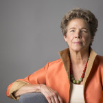Portrait photo of Tia Nelson against a gray backround. She is wearing an orange blazer with a high collar and a green and red beaded necklace