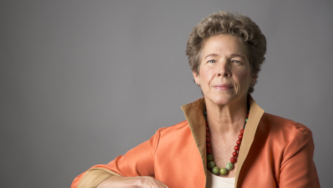 Portrait photo of Tia Nelson against a gray backround. She is wearing an orange blazer with a high collar and a green and red beaded necklace