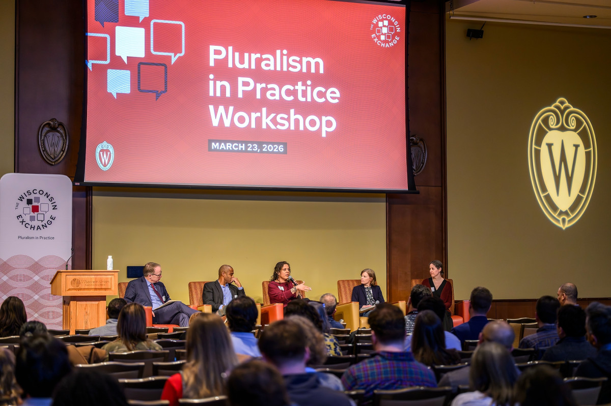 A panel discussion in a lecture hall. Above head, with a large screen with a slide reading "Pluralism in Practice Workshop"