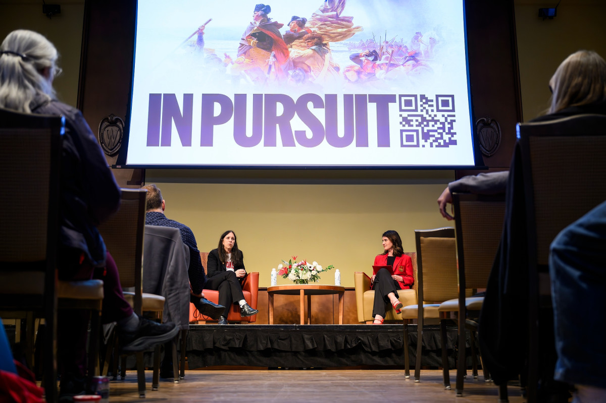 Two panelist talking with a presentation slide above reading "In Pursuit" while an audience observes
