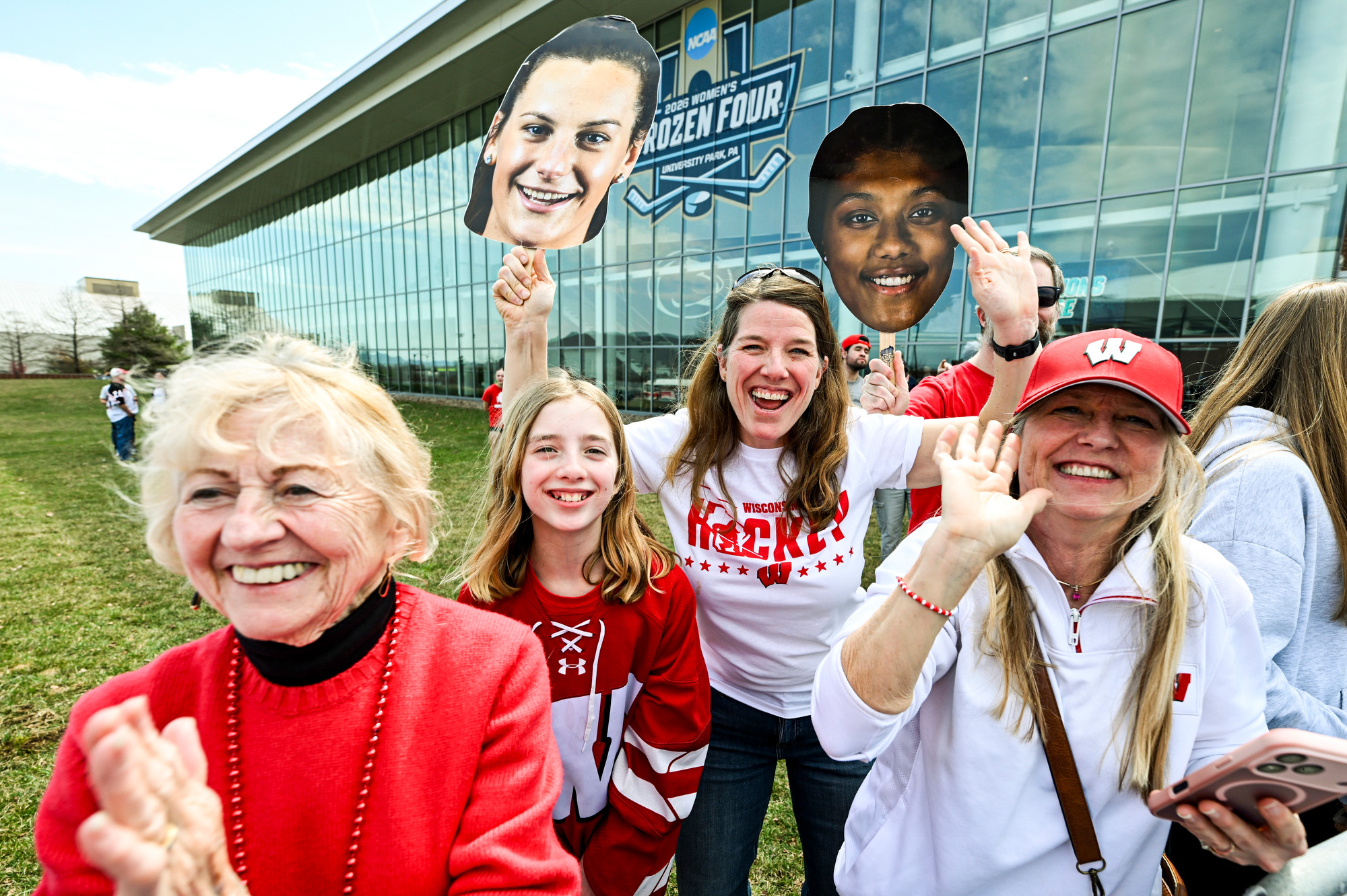Holding face cutouts of members of the Badger women’s ice hockey team, a group of Badger fans stand outside an ice hockey arena and cheer the arrival of the players.