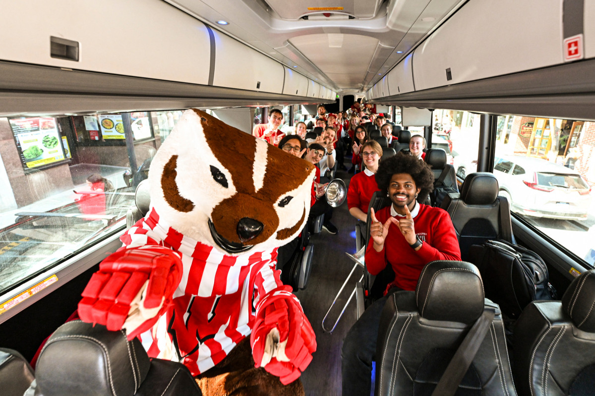 A photo taken from the front of a coach bus shows mascot Bucky Badger and members of the UW Marching Band sitting in their seats and making W signs with their hands