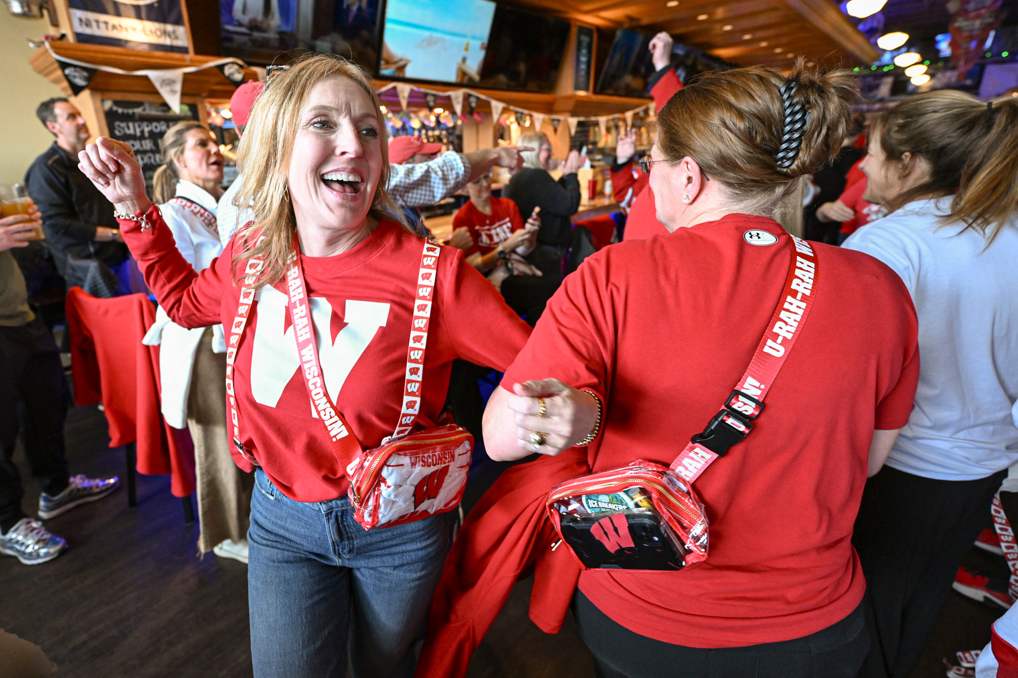 Two women wearing Wisconsin Badger attire link arms and dance the polka inside of a crowded restaurant