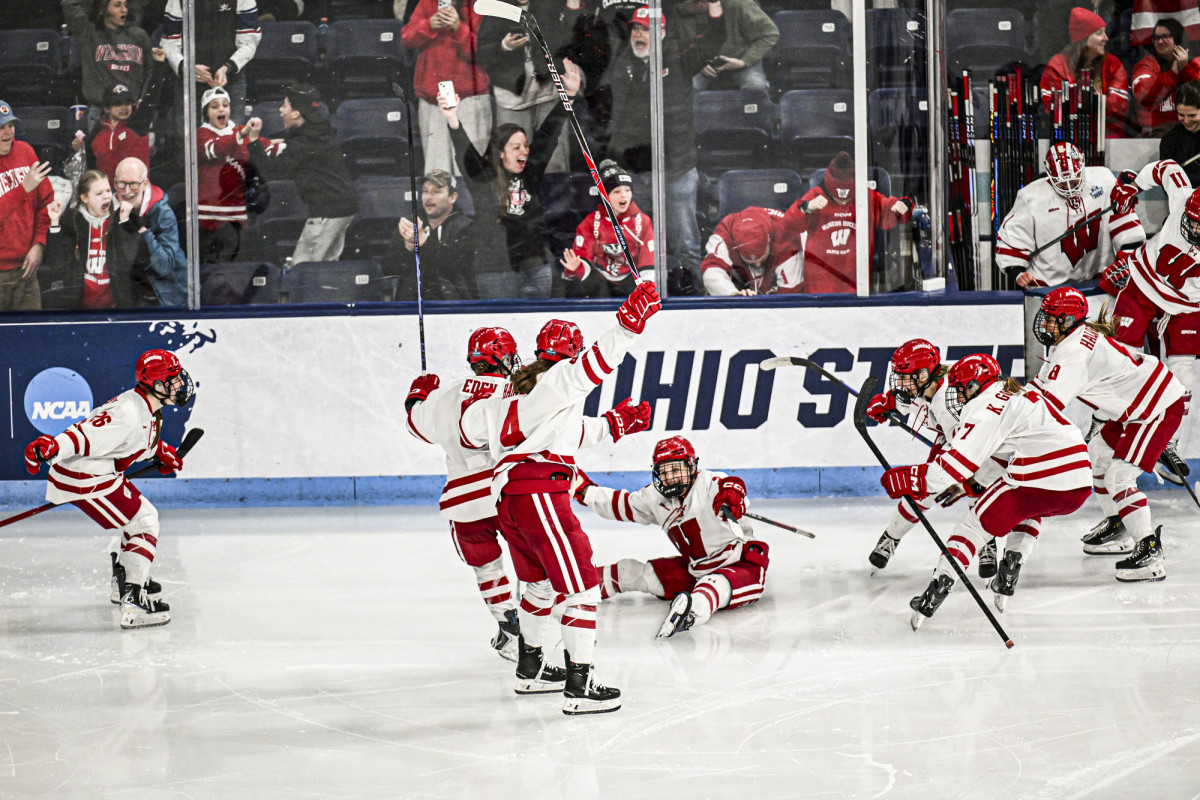 Player Kirsten Simms sits on the ice after scoring the winning goal as fellow Badger players rush to celebrate her shot.