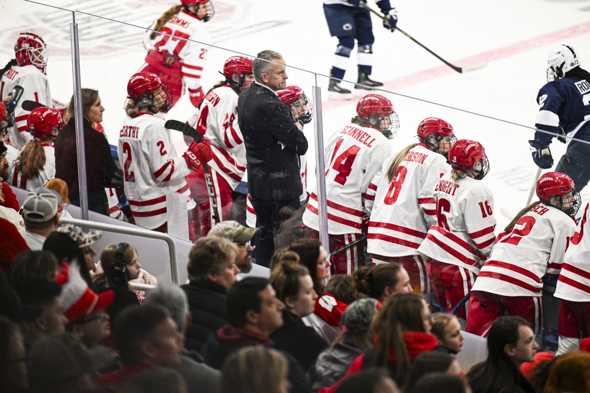 Members of the Badger women’s hockey team sit on the bench and watch the action on the ice along with Coach Mark Johnson who stands behind them with arms crossed and observes.