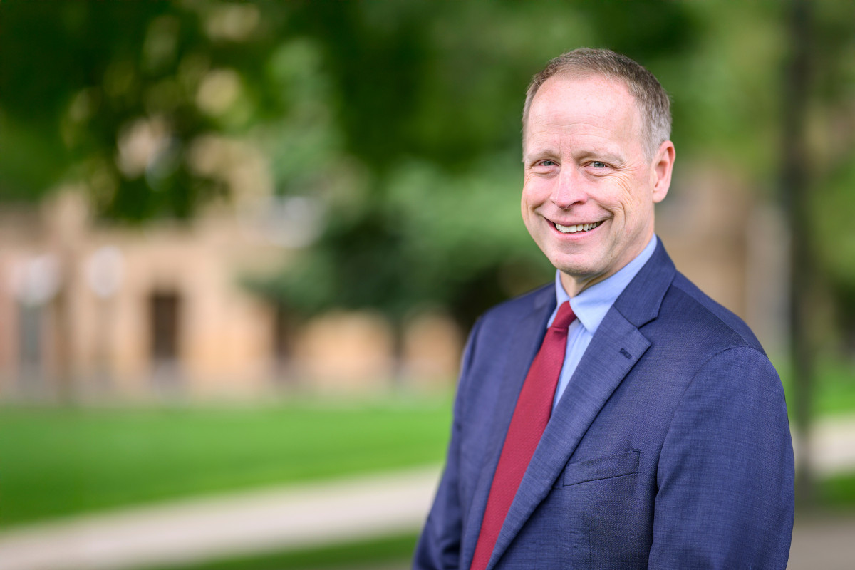 Portrait photo of John Zumbrunnen smiling. He is wearing a blue suit and red tie and standing outdoors in front of a blurred background of green trees.