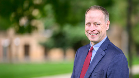 Portrait photo of John Zumbrunnen smiling. He is wearing a blue suit and red tie and standing outdoors in front of a blurred background of green trees.