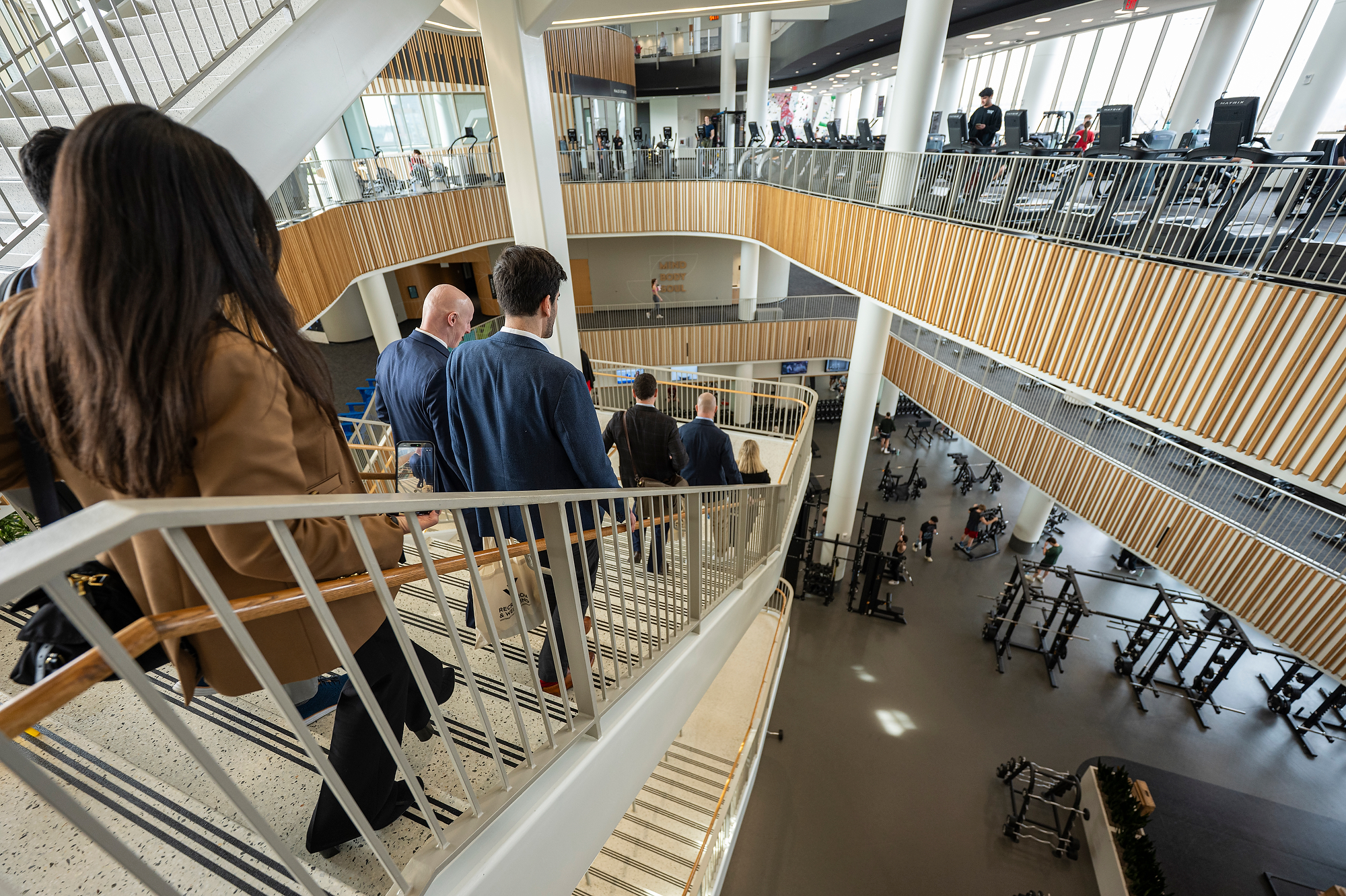 A group of people walk down a wide interior staircase overlooking multiple floors of an open, modern facility with fitness equipment visible below.