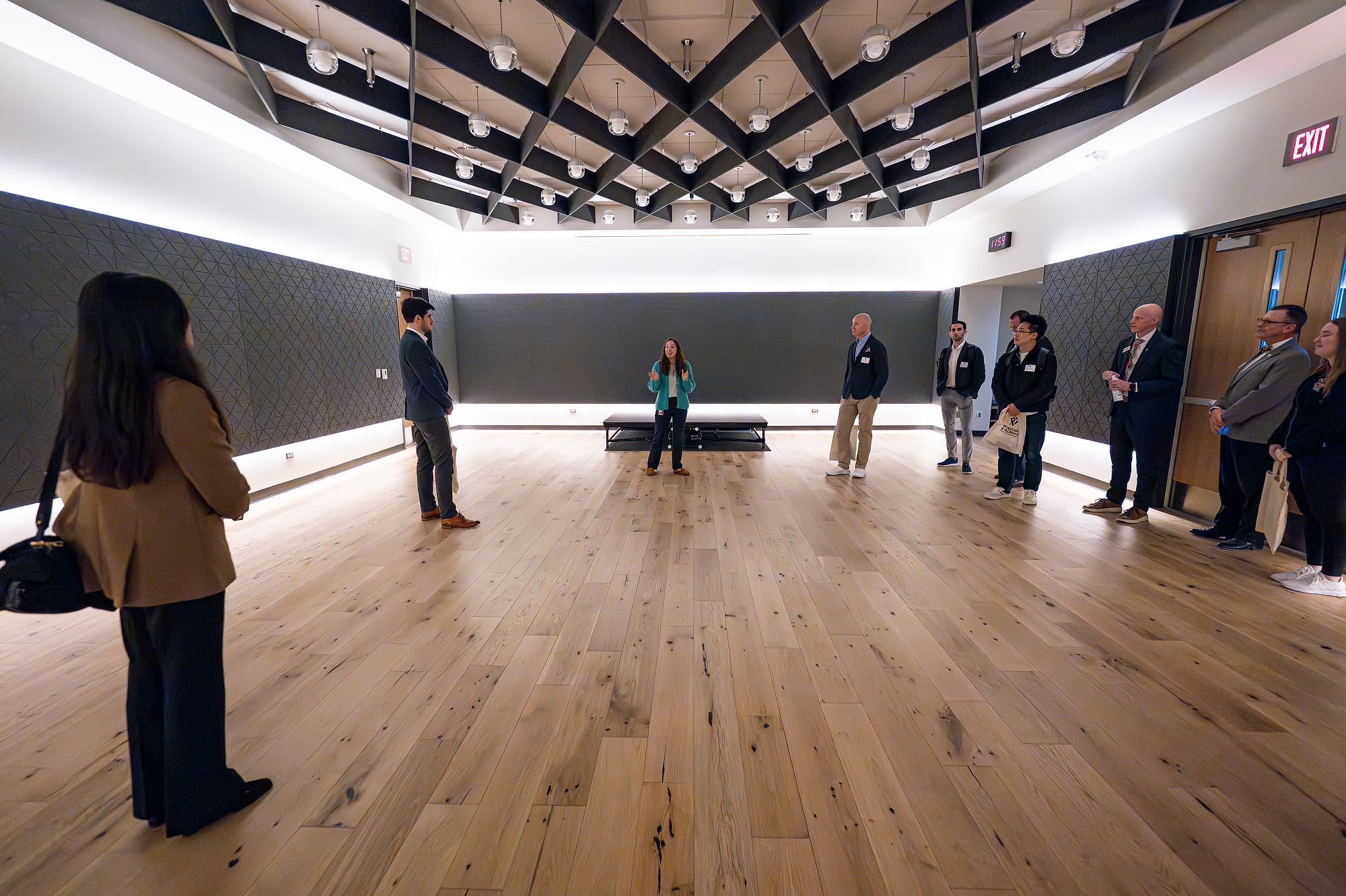A group of people stand spaced around a large wood‑floored room while a speaker at the front provides an explanation during a tour.