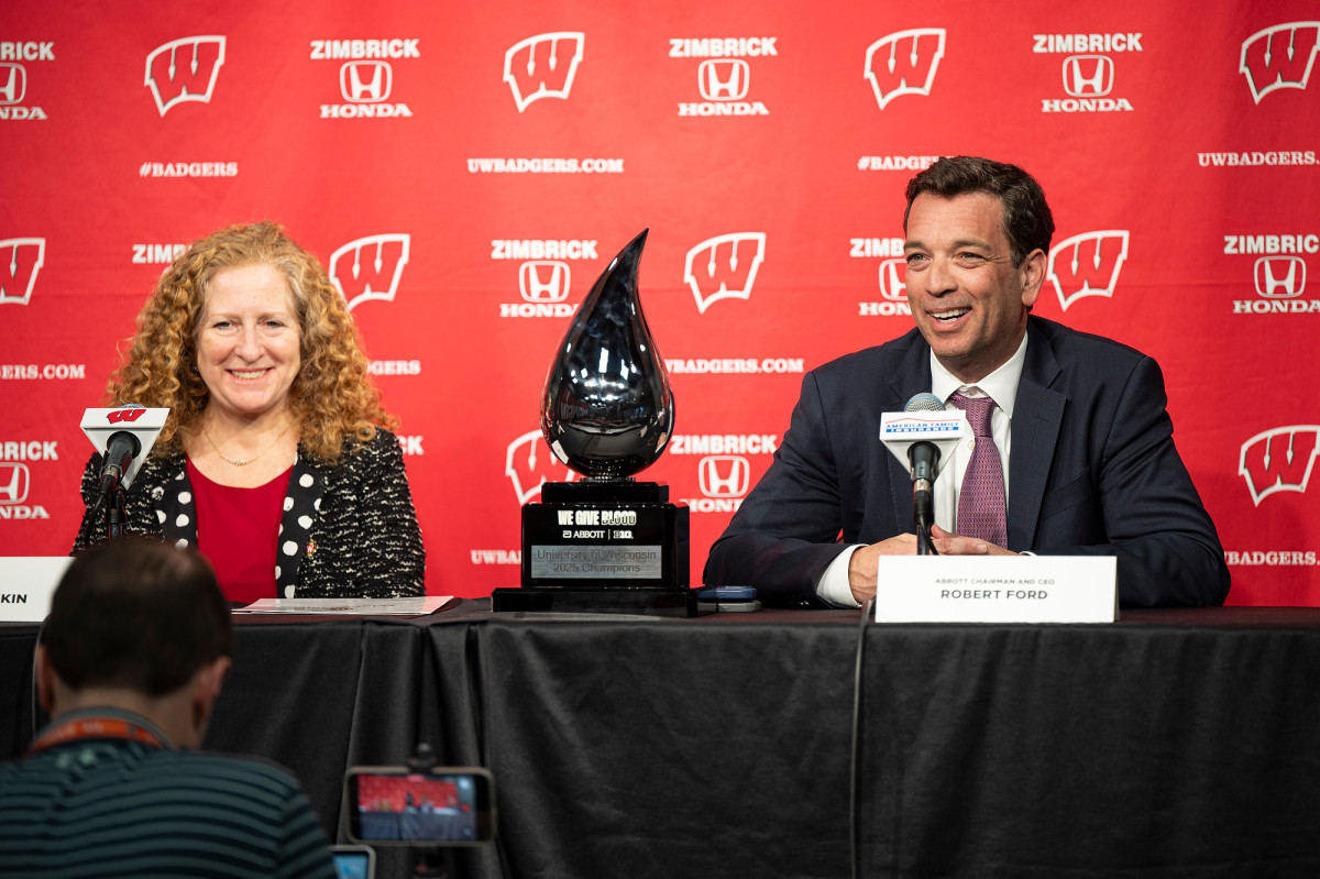 Two speakers sit at a press‑conference table with microphones in front of a red backdrop, with a glass award in the shape of a drop of blood displayed between them.