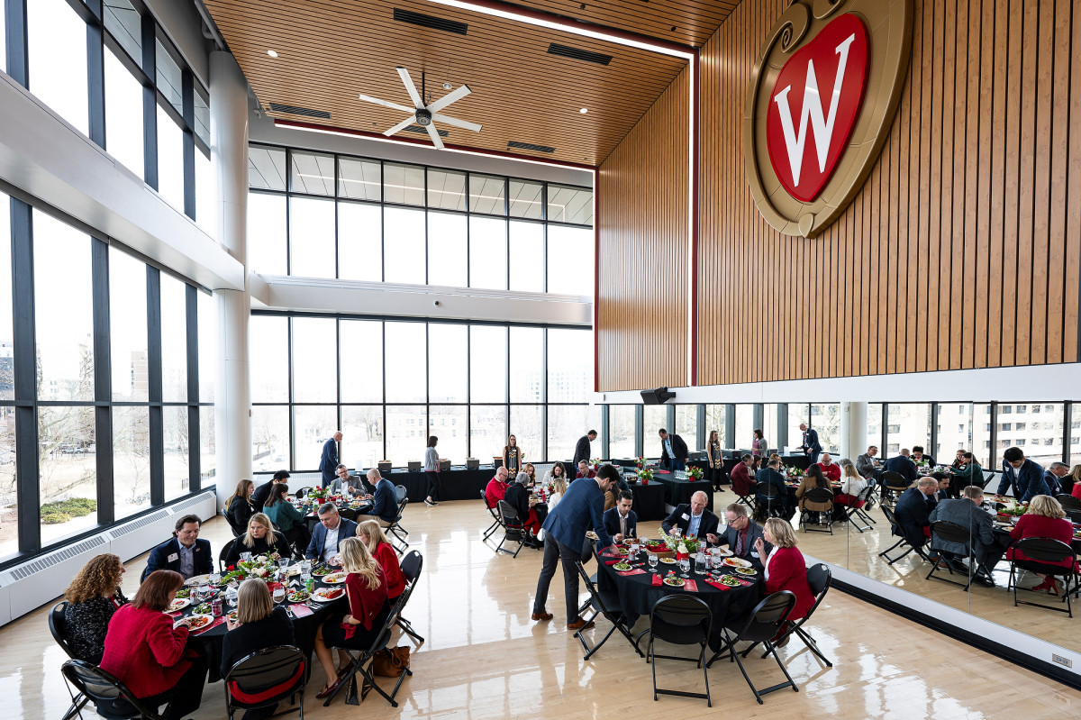 People seated at round tables enjoy a meal in a large, bright room in with floor‑to‑ceiling windows and a prominent ‘W’ crest on a wood-paneled wall.
