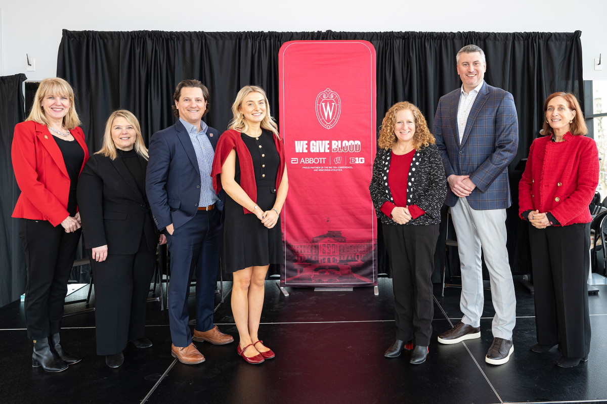 A group of people stand on a stage in front of a backdrop with University of Wisconsin and blood donation-themed graphics.