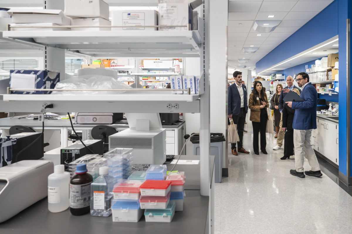 A group of people stand in a bright research laboratory as a staff member speaks to them, with shelves of lab supplies and equipment visible in the foreground.