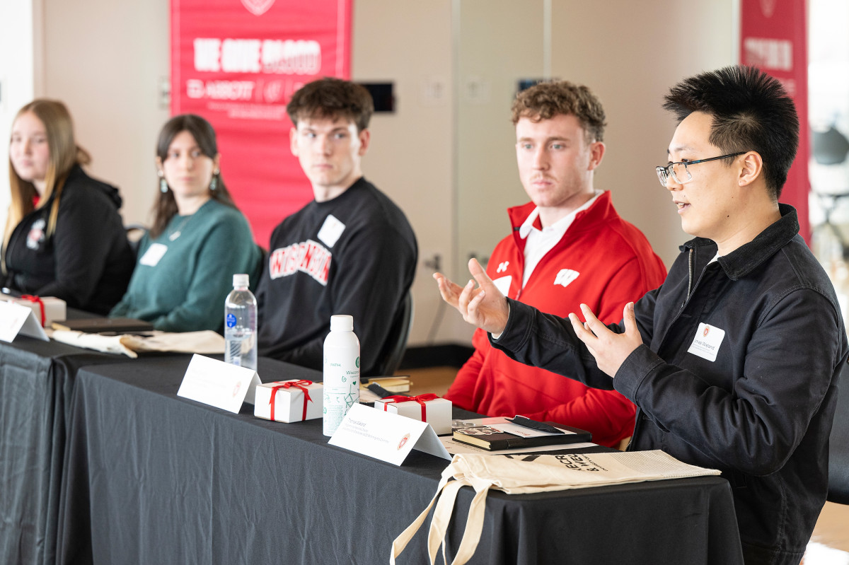A panel of student participants sits at a table with name cards and materials while one person speaks and gestures with both hands during a discussion.