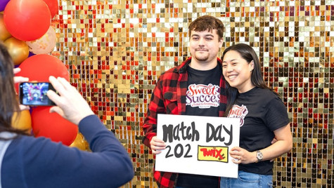 Two people stand in front of a gold, glittering backdrop while holding up a sign for Match Day.