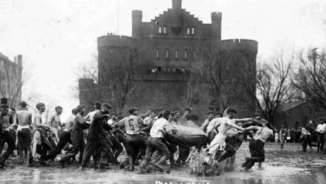A historic black‑and‑white photograph of a group of young men engaged in a muddy game with a large sack in rain‑soaked field Library Mall. The Red Gym is in the background.