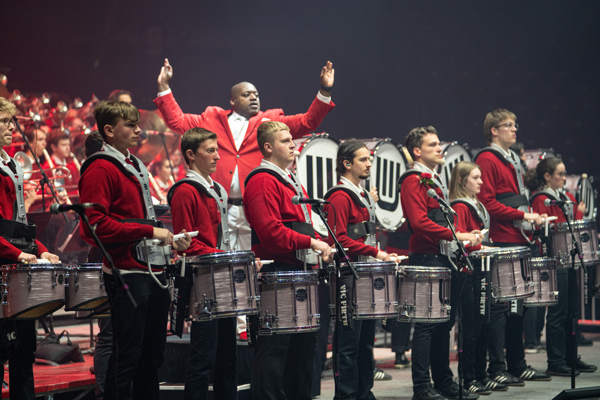 A drum line in front of a larger band, with the conductor raising his hands behind them.