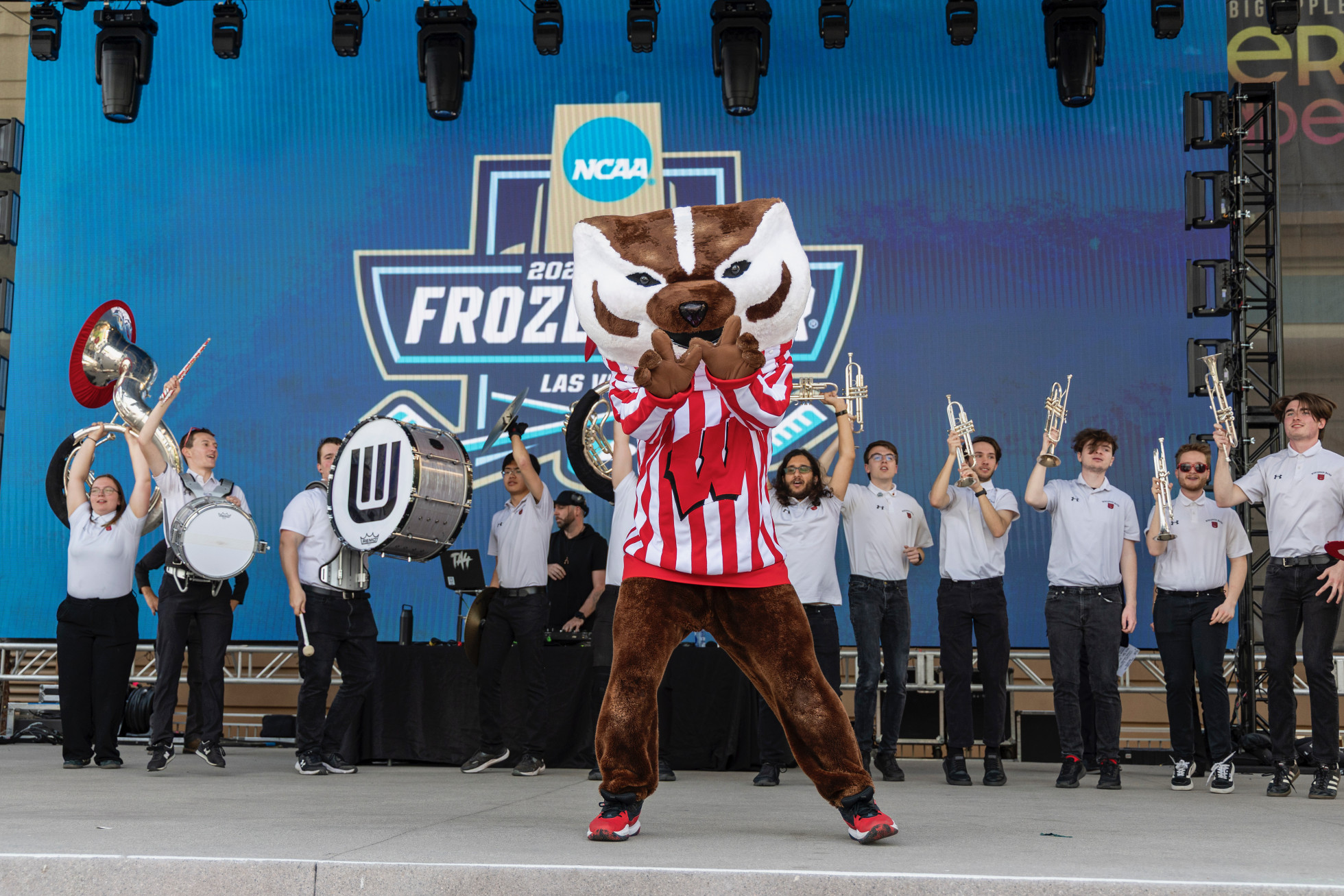Bucky Badger stands on a stage making a W sign with his hands. Behind him are members of the UW marching band holding their brass and percussion instruments high. At the back of the stage is a banner that says NCAA 2026 Frozen Four Las Vegas.