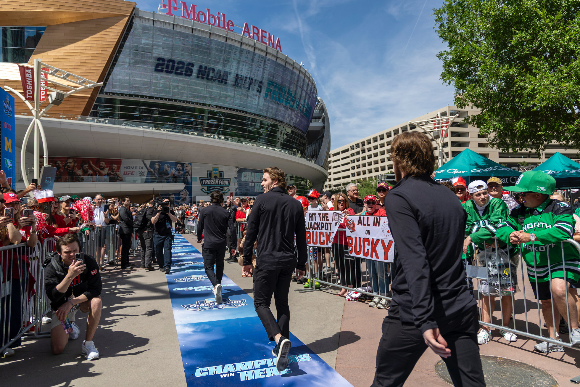 Three members of the Wisconsin men's hockey team walk away from the camera through a crowd of cheering fans to enter the T Mobile Arena.