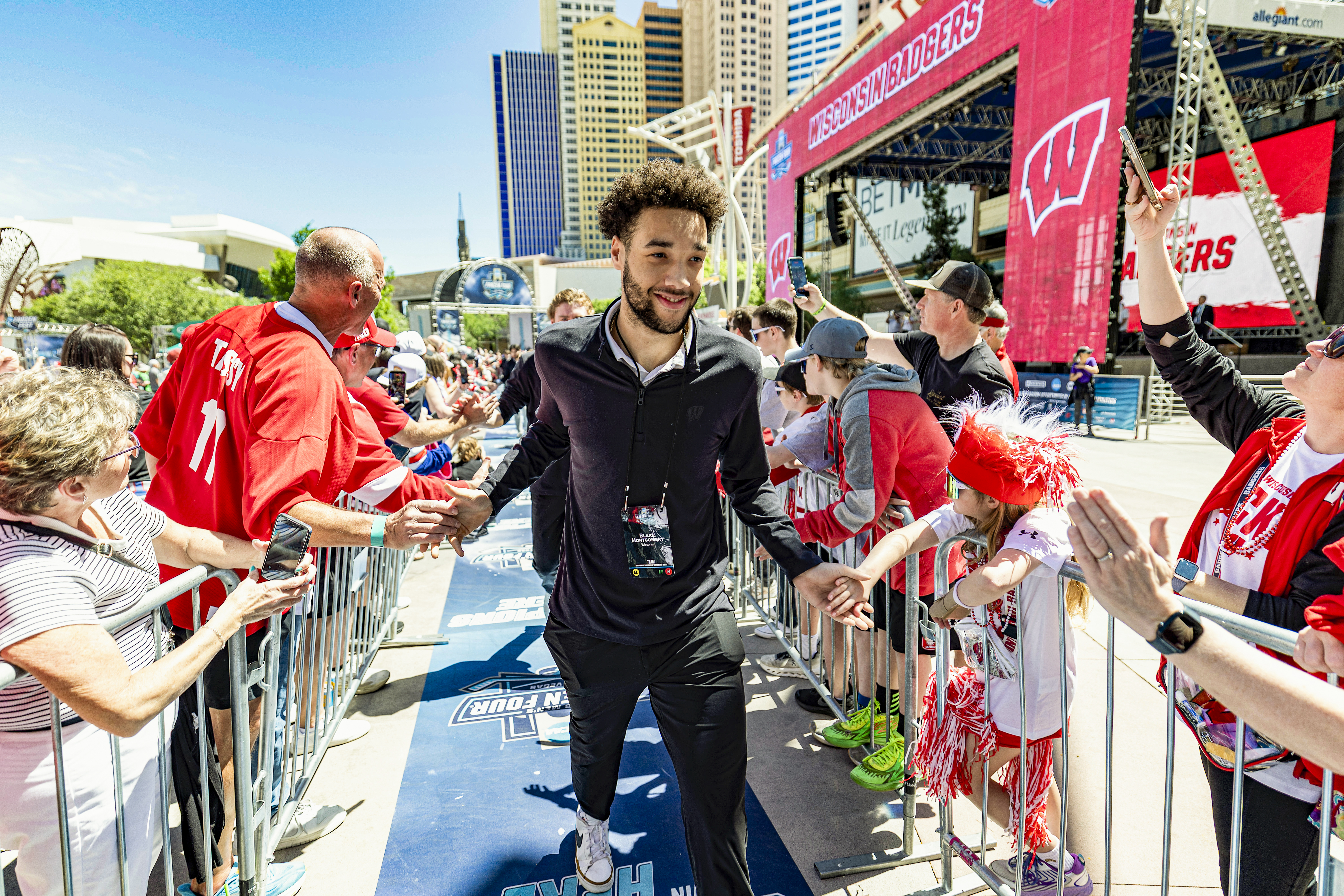A man wearing black track suits give high fives as they walk through a crowd of cheering UW fans. Behind them is a grand stand with the words Wisconsin Badgers and the Las Vegas skyline.