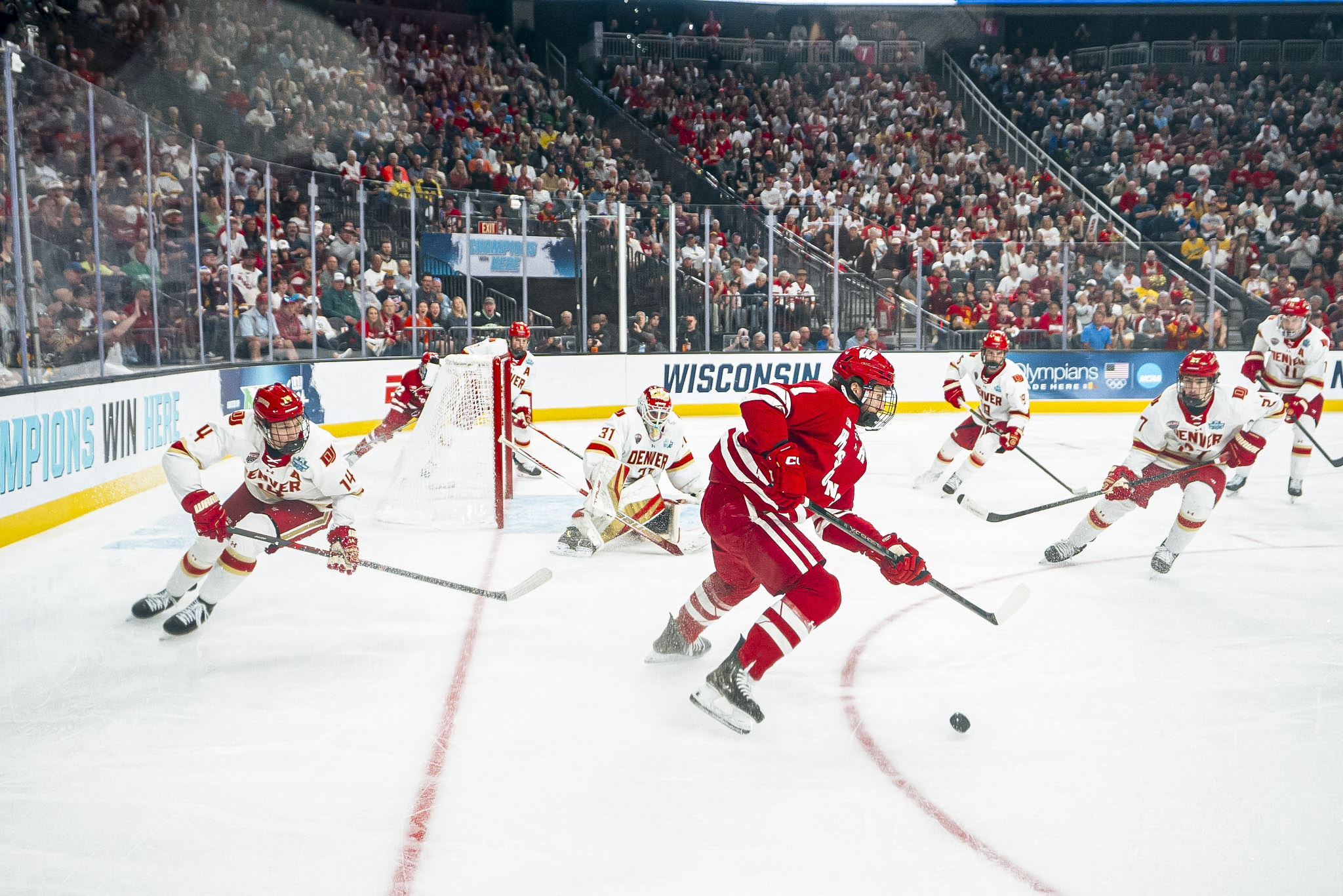 Six Denver players wearing white try to defend their goal against one Wisconsin player in red. Wisconsin's player has the pick and is looking to pass to a teammate.