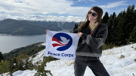 A Peace Corps volunteer holds up a banner with the Peace Corps logo on a snowy summit.