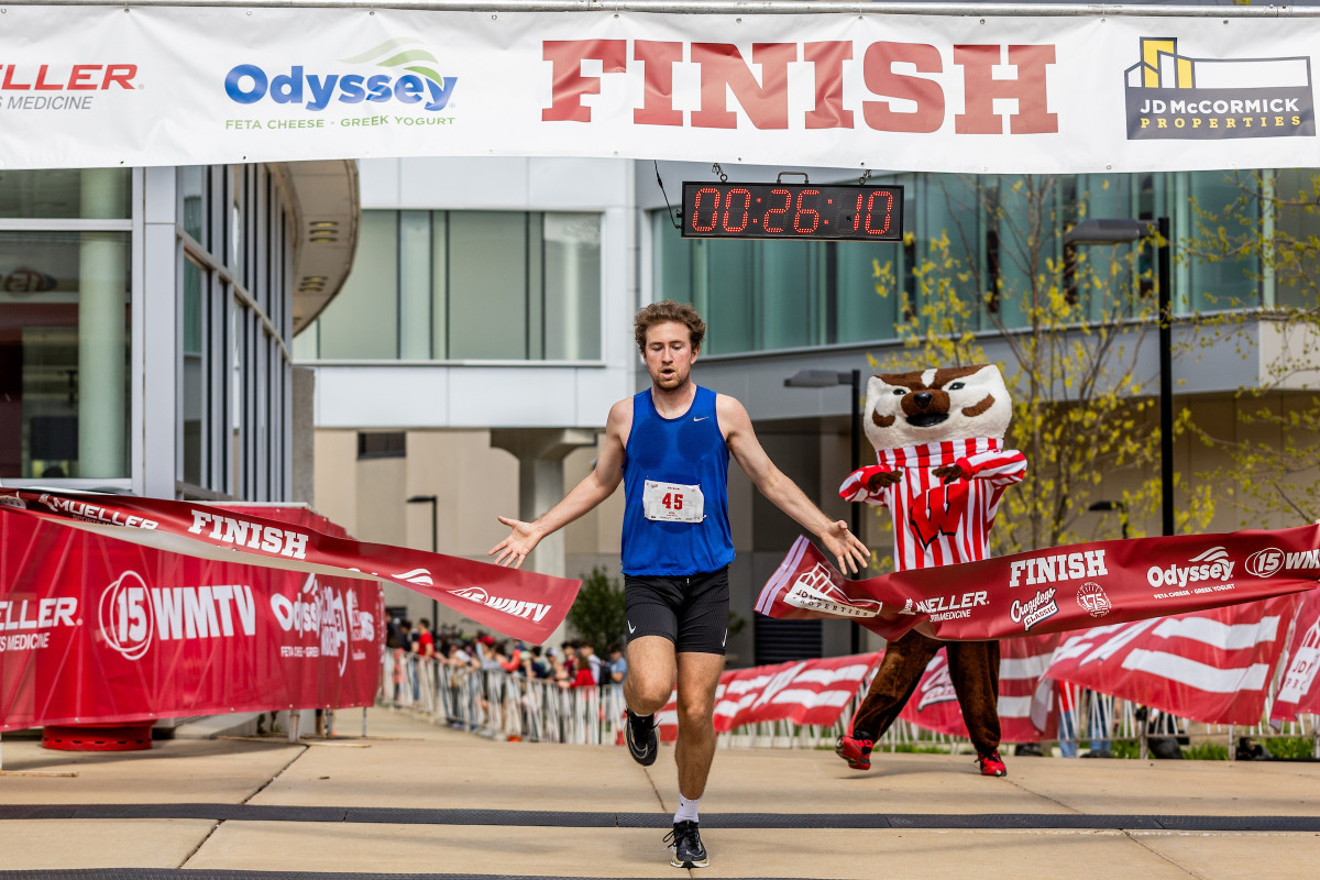 A runner crosses a finish line ribbon with Bucky Badger applauding in the background.