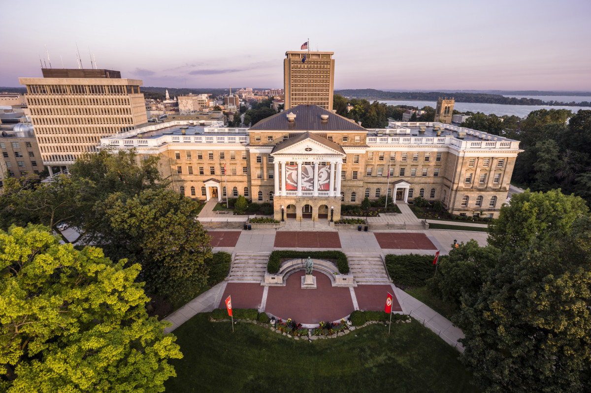 An aerial view of an academic building, Bascom Hall, at the University of Wisconsin–Madison. There is foliage on trees and a green lawn. It is dawn so the sky is light pink, blue and purple.