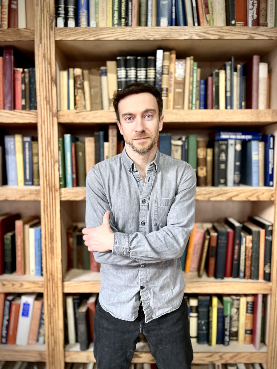 Author Eliot Stein stands with arms crossed in front of wooden shelves filled with books