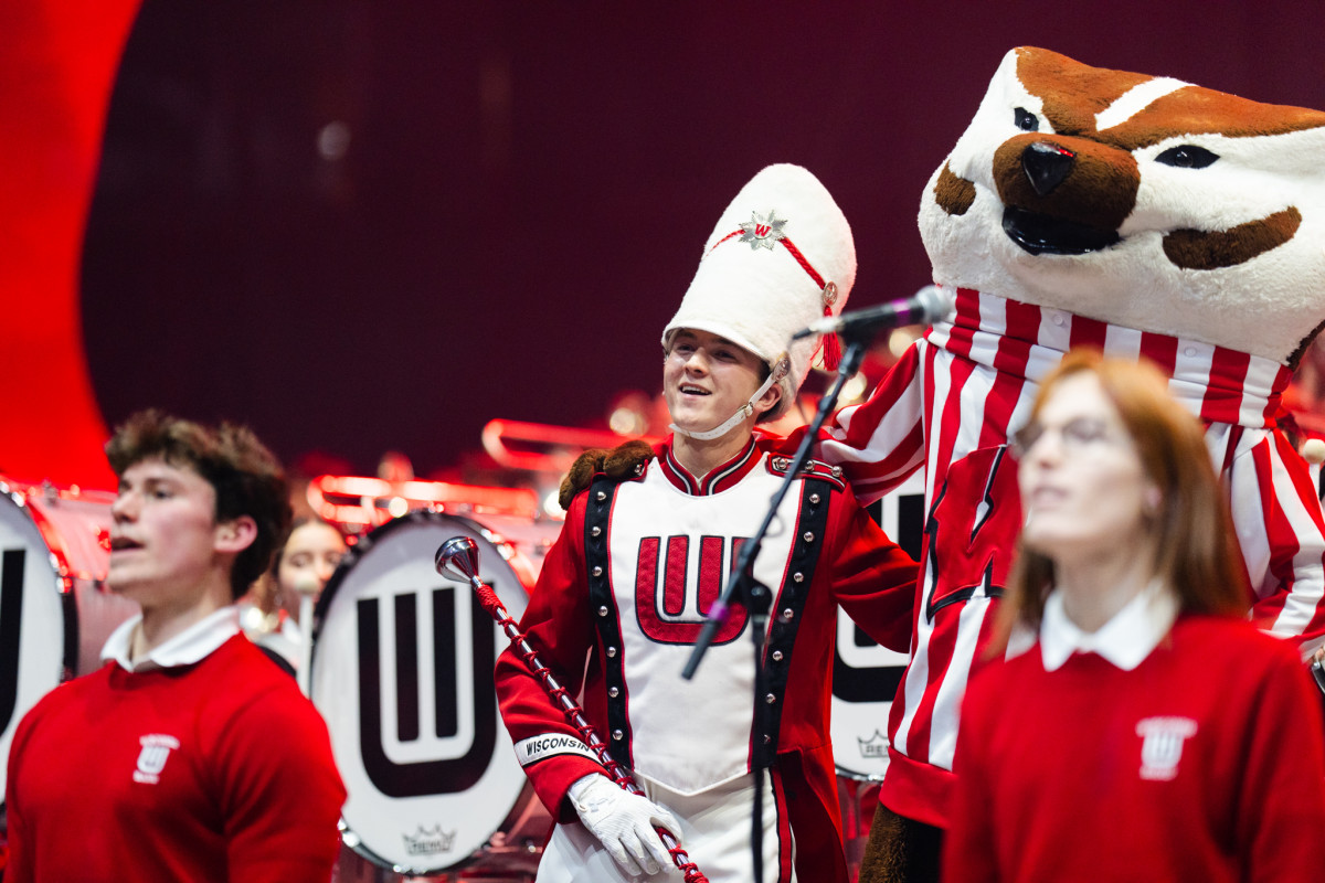 A drum major for the UW Marching Band wraps his arm around Bucky the Badger mascot in a concert venue.