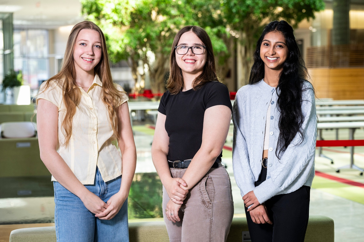 Three students stand and smile for a portrait photo in the brightly lit atrium of a building.