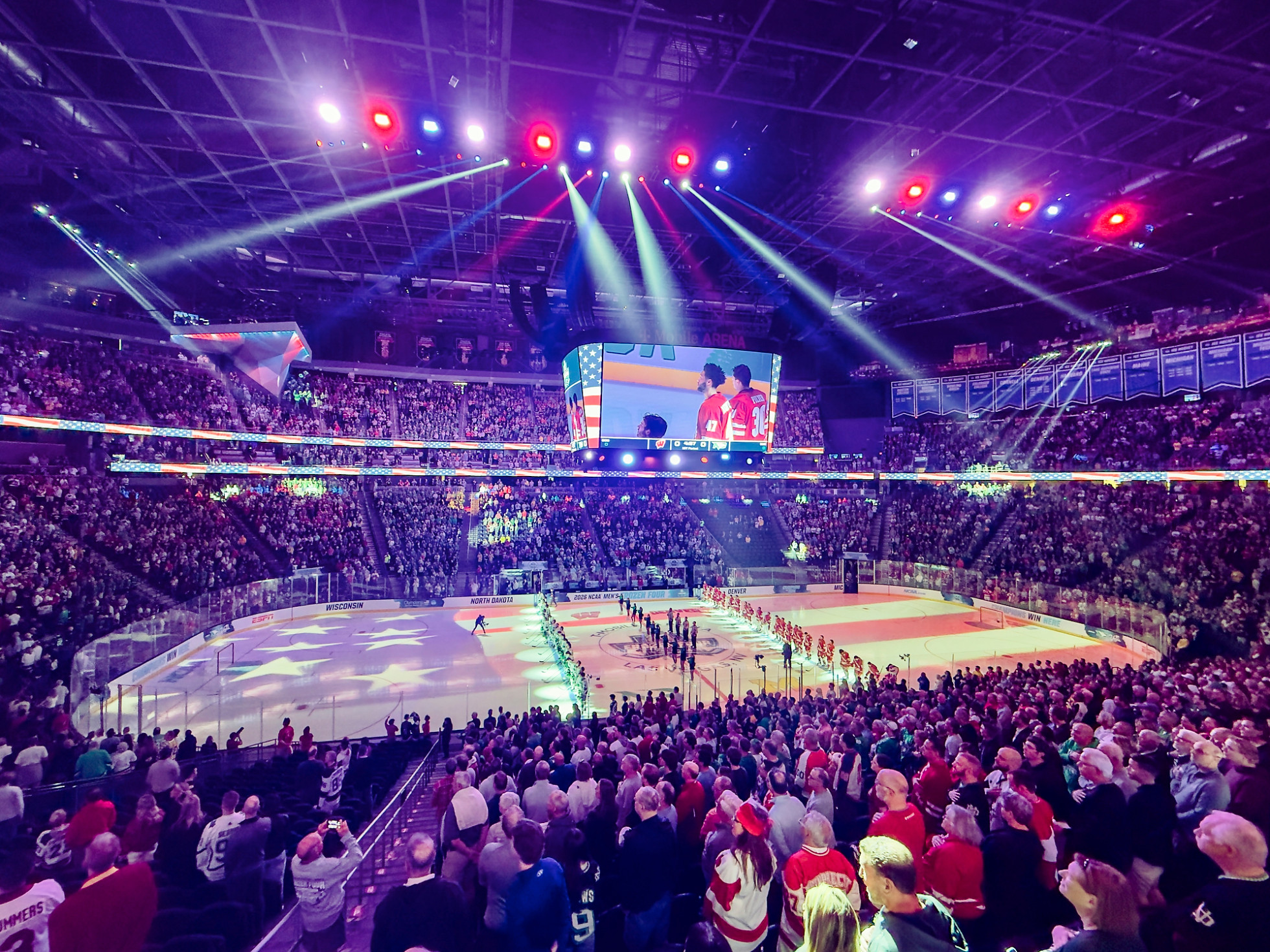 In an ice hockey arena, the two teams stand in a line, and the crowd is on its feet for the national anthem. Lights from the rafters shine down in red, white and blue to paint an American flag on the ice.