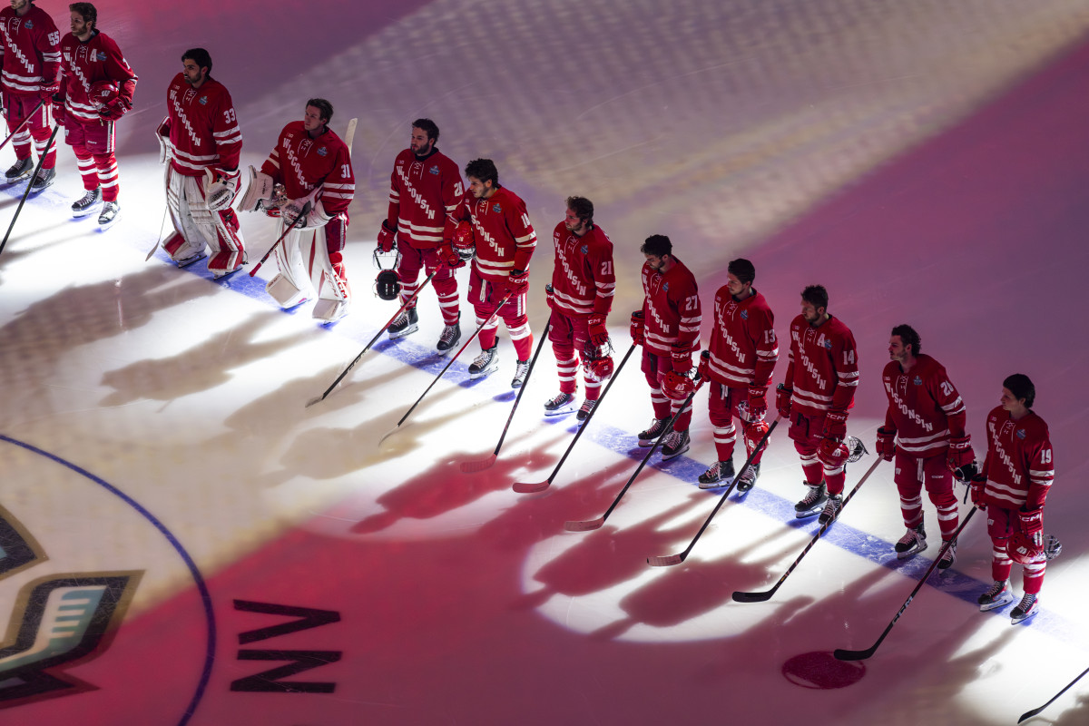 Seen from above, a line of hockey players wearing red UW–Madison uniforms stand at center ice for the national anthem.