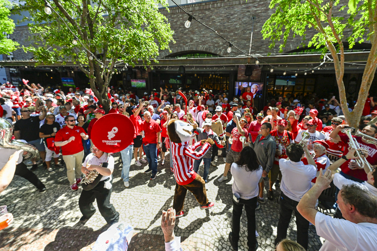 A crowd of people wearing red and white gather on a patio outside a low brick building on a sunny day. They're dancing with Bucky Badger while a member of the UW marching band plays the tuba.