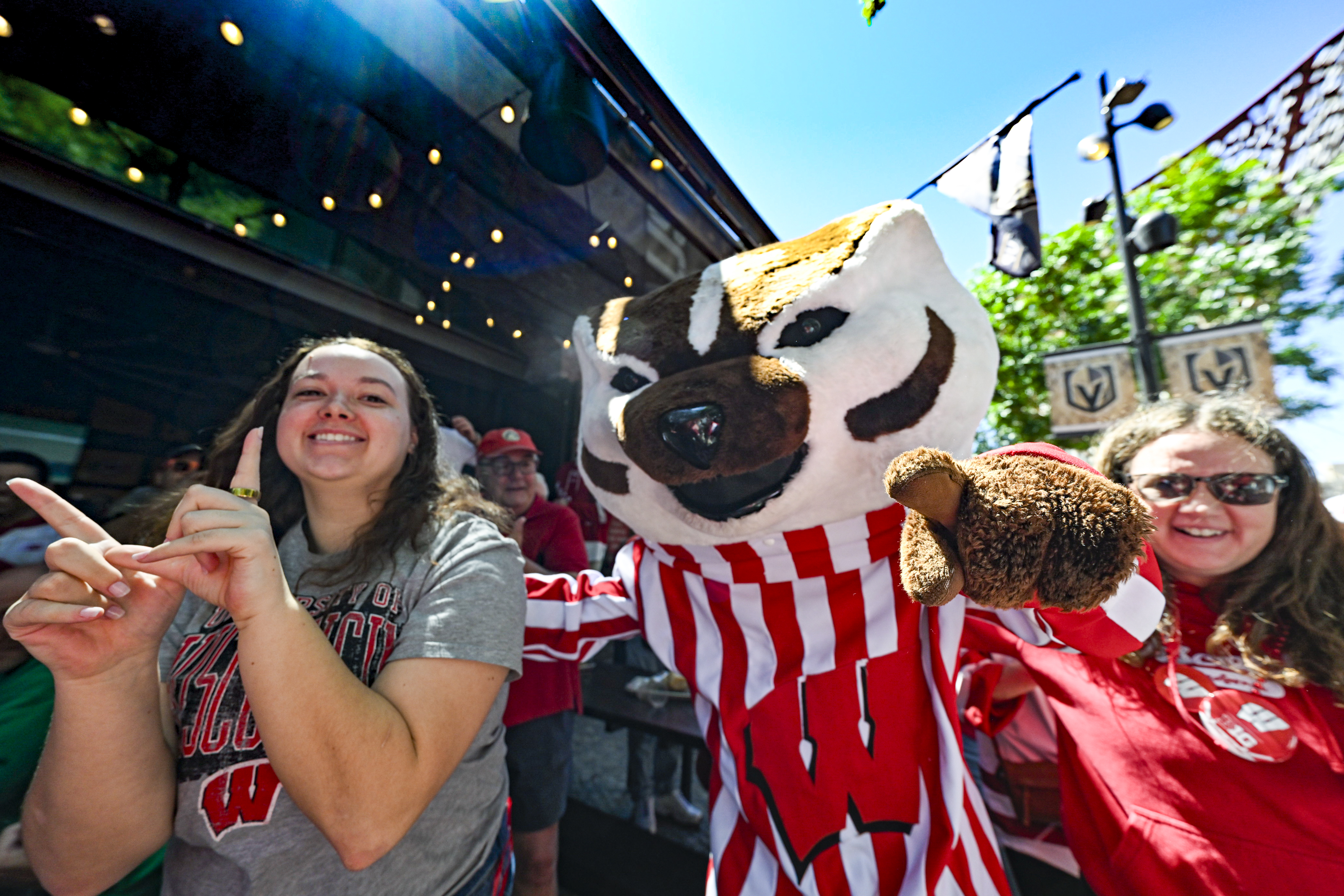 Two Wisconsin fans wearing UW–Madison T-shirts stand outdoors on either side of Bucky Badger. Both fans are smiling and Bucky is pointing directly to the camera.