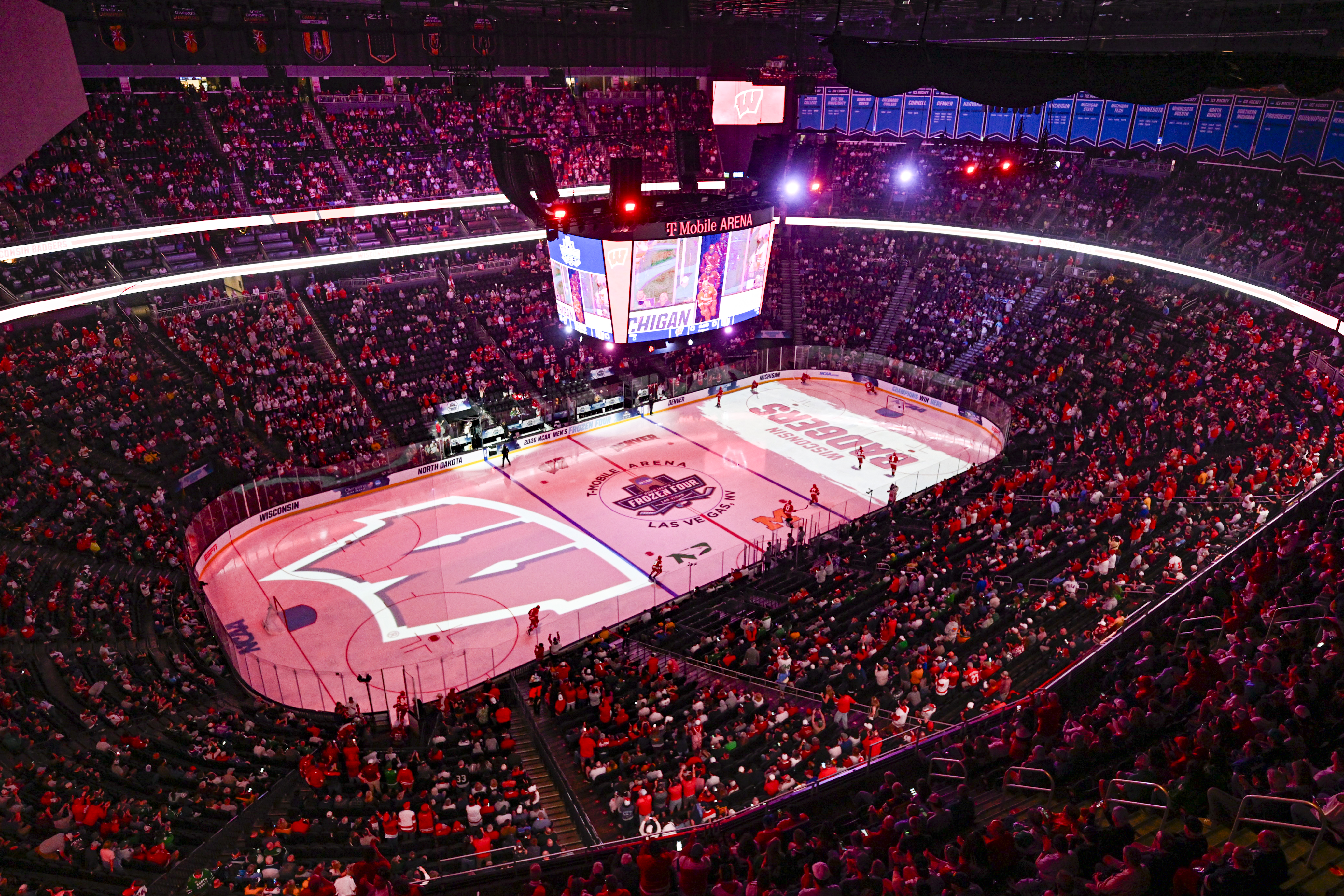 A wide view of a crowd-filled hockey arena with the lights dimmed while players begin to skate onto the ice. Projected on the ice are UW–Madison's W logo and the words Wisconsin Badgers.