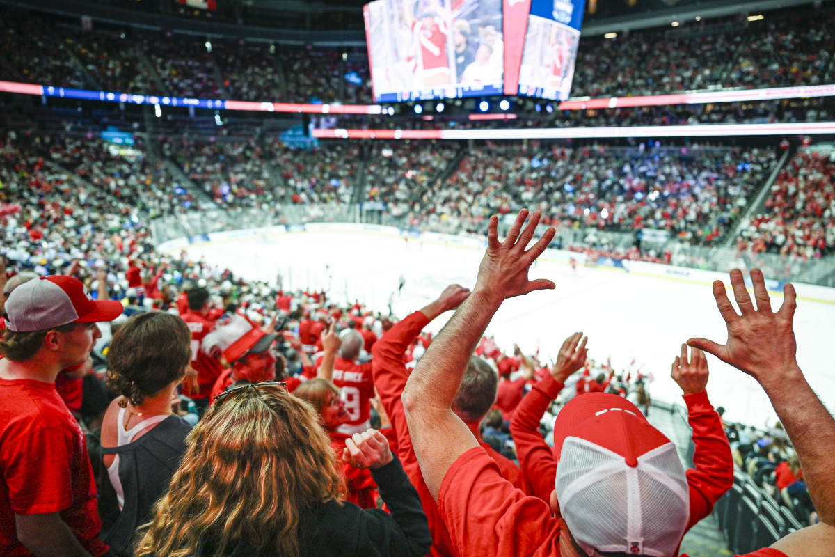 Looking out at the ice rink from the back of the stadium, a sea of Wisconsin fans in red and white stand with their hands raised in excitement.