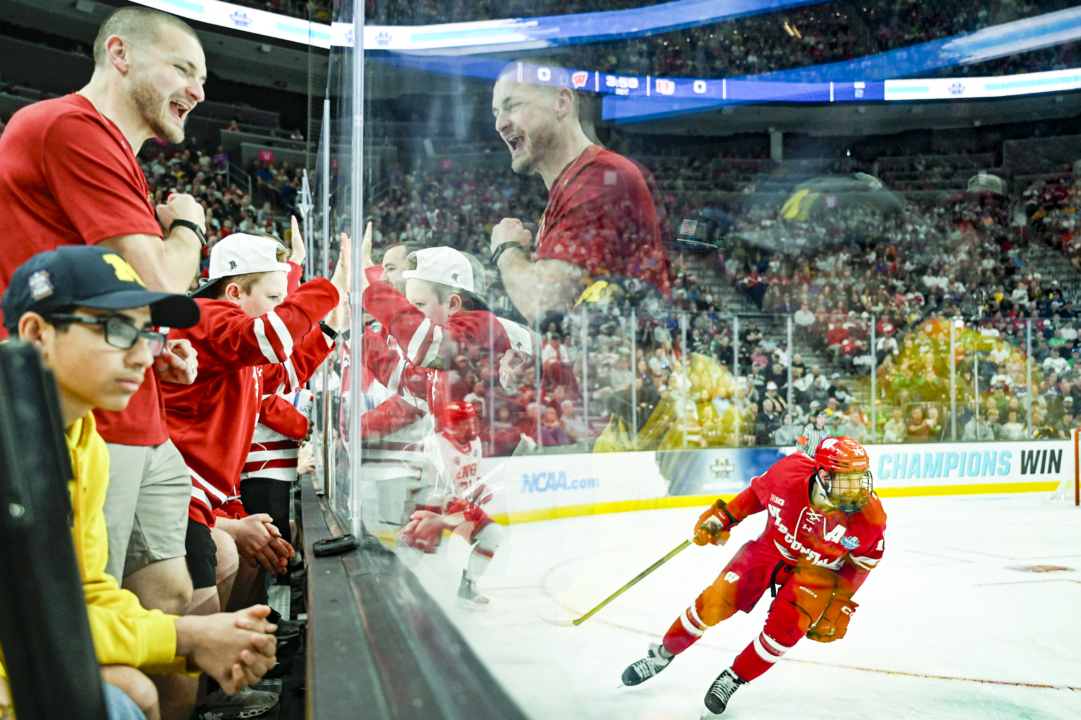 Wisconsin fans in red and white stand and cheer as they look through the glass to watch a Wisconsin player in red skate by.