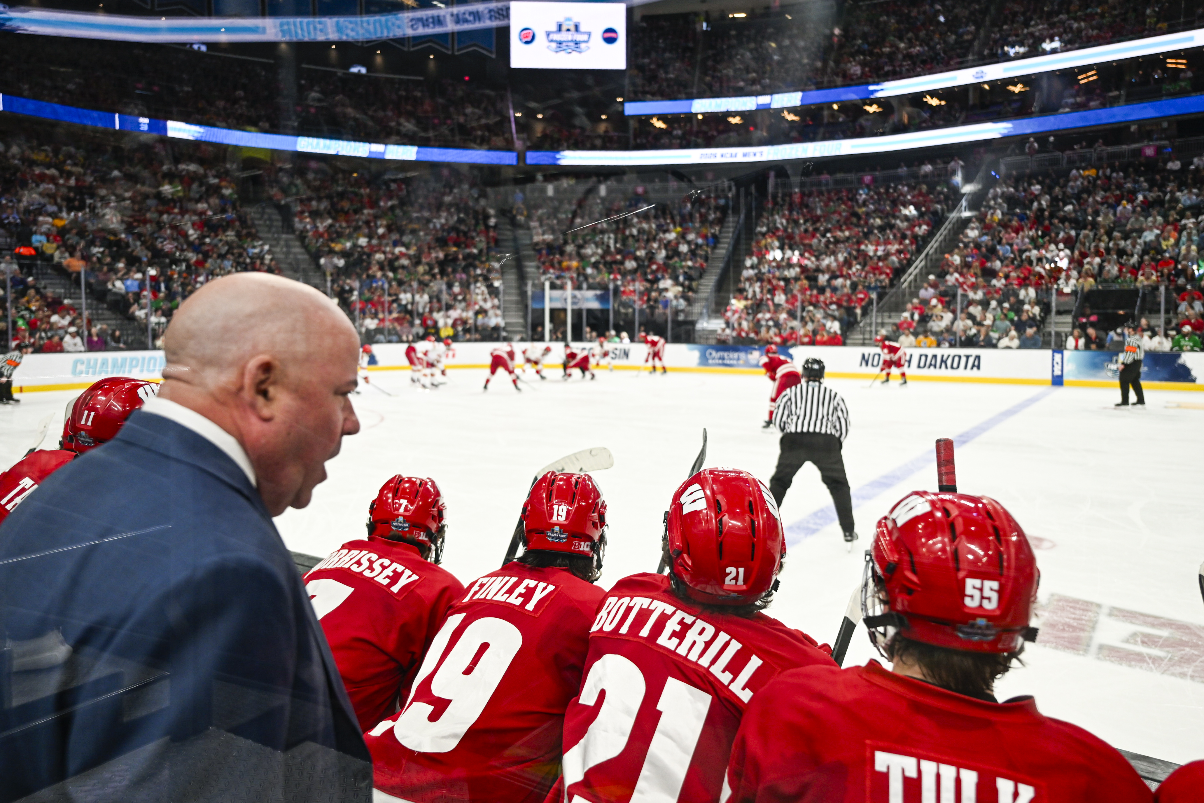 A man wearing a suit speaks to players in the box as they look out on the ice to watch the game.