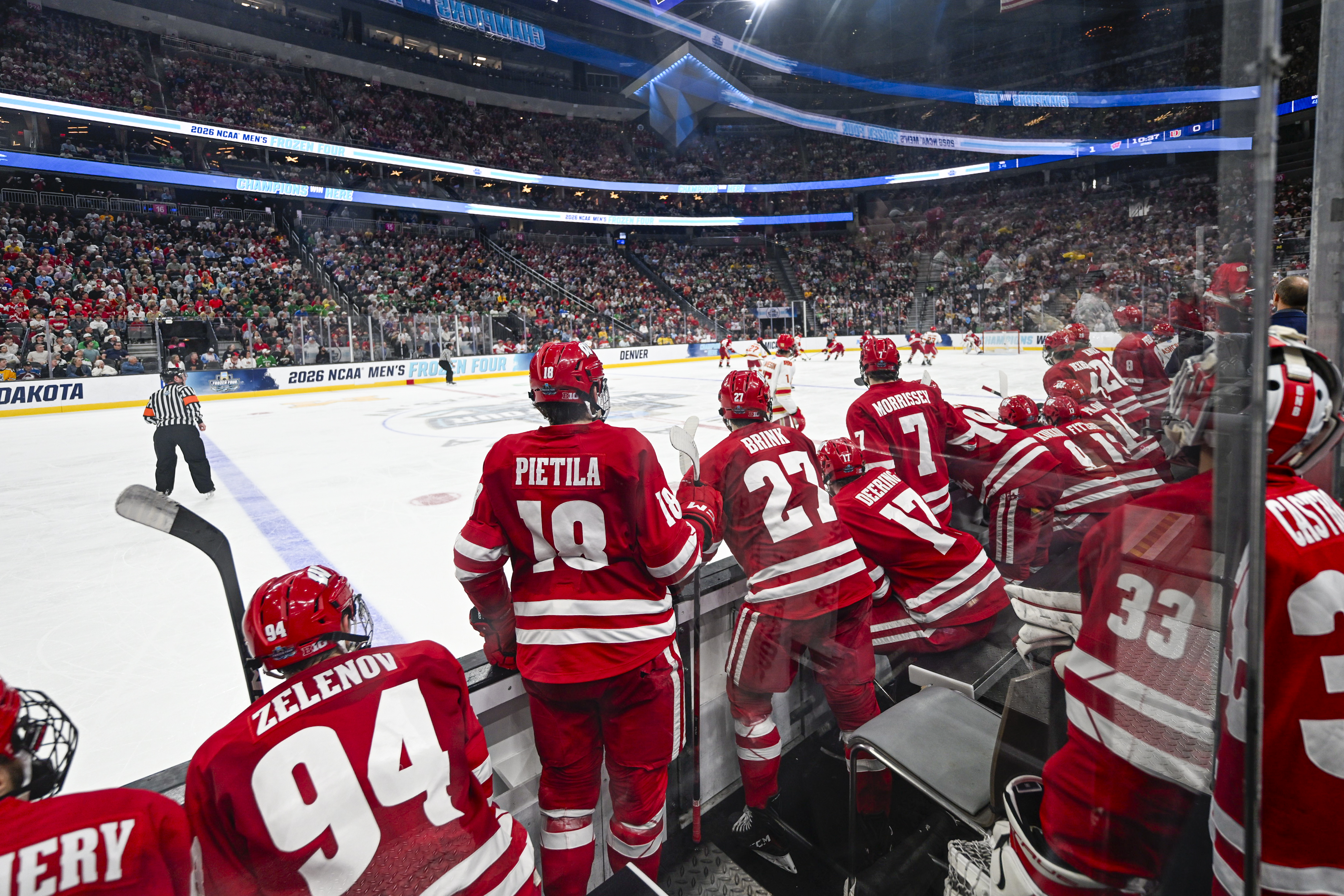 A group of Wisconsin players stand in the box on the side of the rink watching the game.
