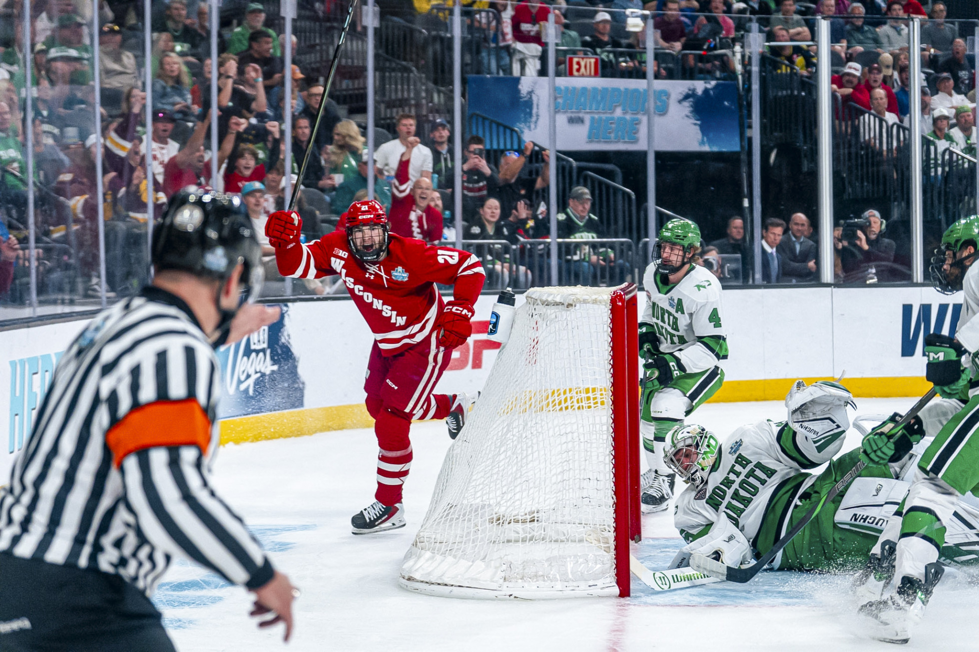 Wisconsin player 21 celebrates his goal with his stick overhead as the North Dakota goalie falls to the ice.