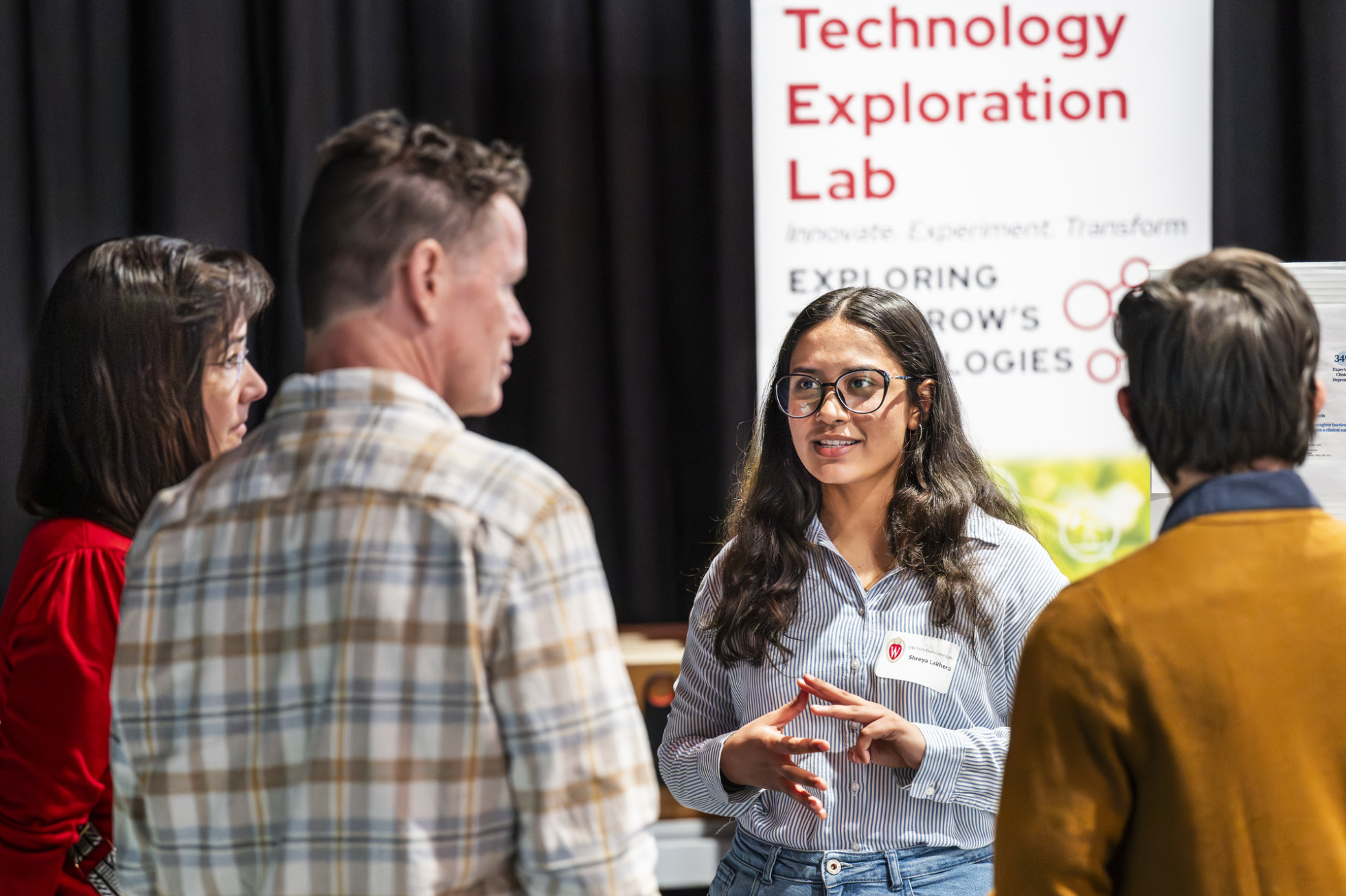 A group of four people stand together talking. Behind them hangs a sign for the Technology Exploration Lab.