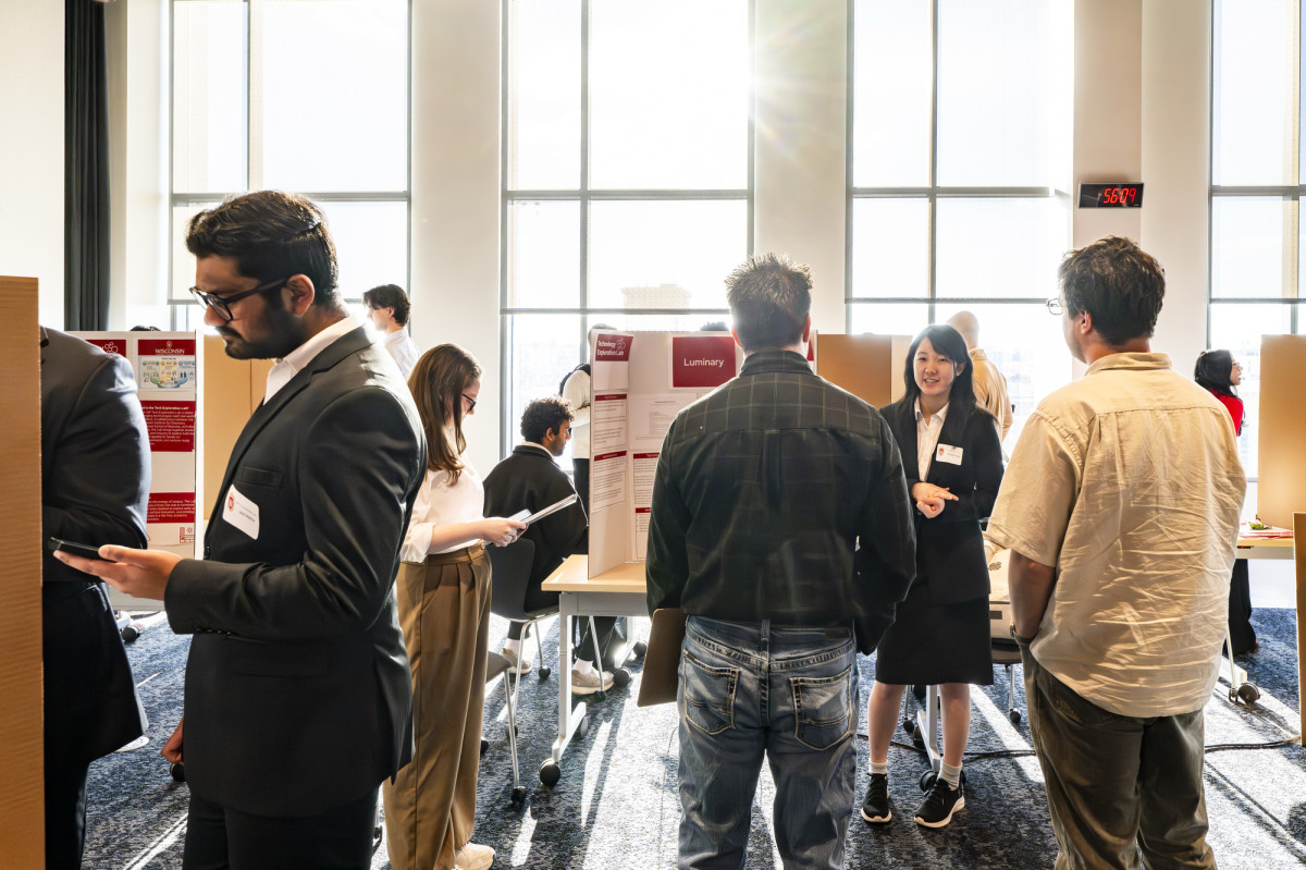 In a brightly lit exhibition hall, students stand by their posters to explain their work as visitors mill through.