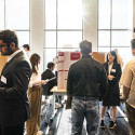 In a brightly lit exhibition hall, students stand by their posters to explain their work as visitors mill through.