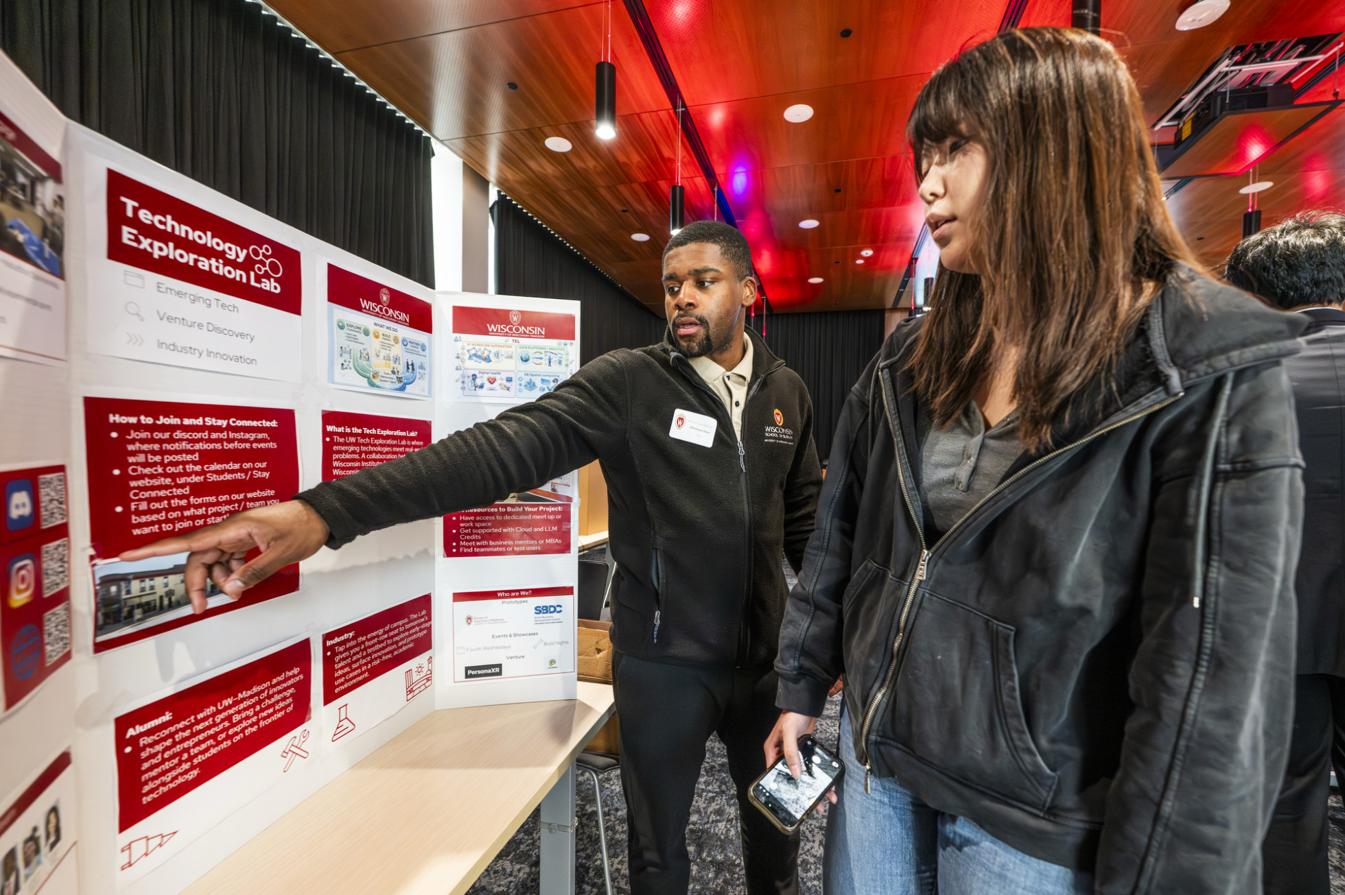 A student points to a research poster as he explains his work to a visitor.