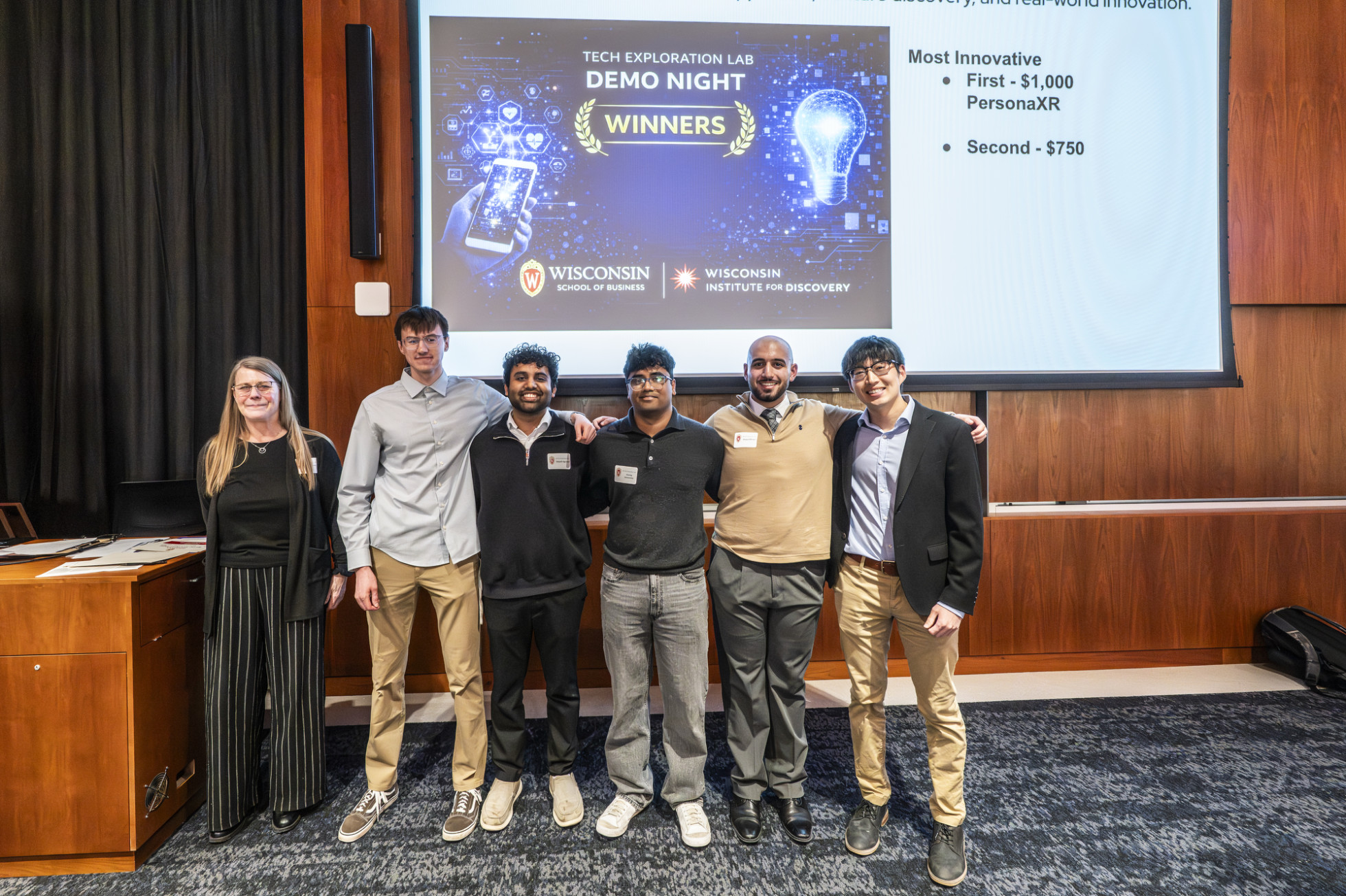 A group of six people smile as they stand arm in arm in front of a projector screen with the words Tech Exploration Lab Demo Night Winners.