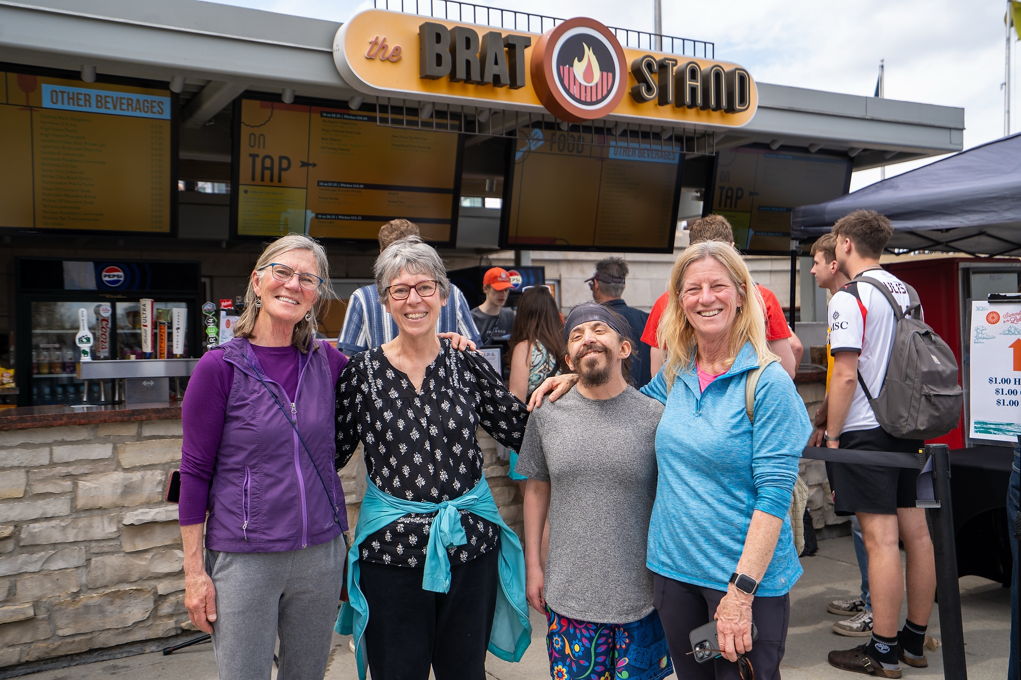 Four people pose with arms wrapped around each other in front of the Brat Stand at Memorial Union. 
